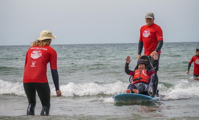 A disabled lady on a seated surfboard on a wave in the sea with her arms in the arm and smiling, with a surf instructor surfing on the board with her